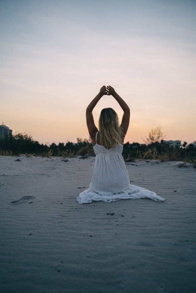 A woman in a white dress meditates on a sandy beach during a serene sunset.